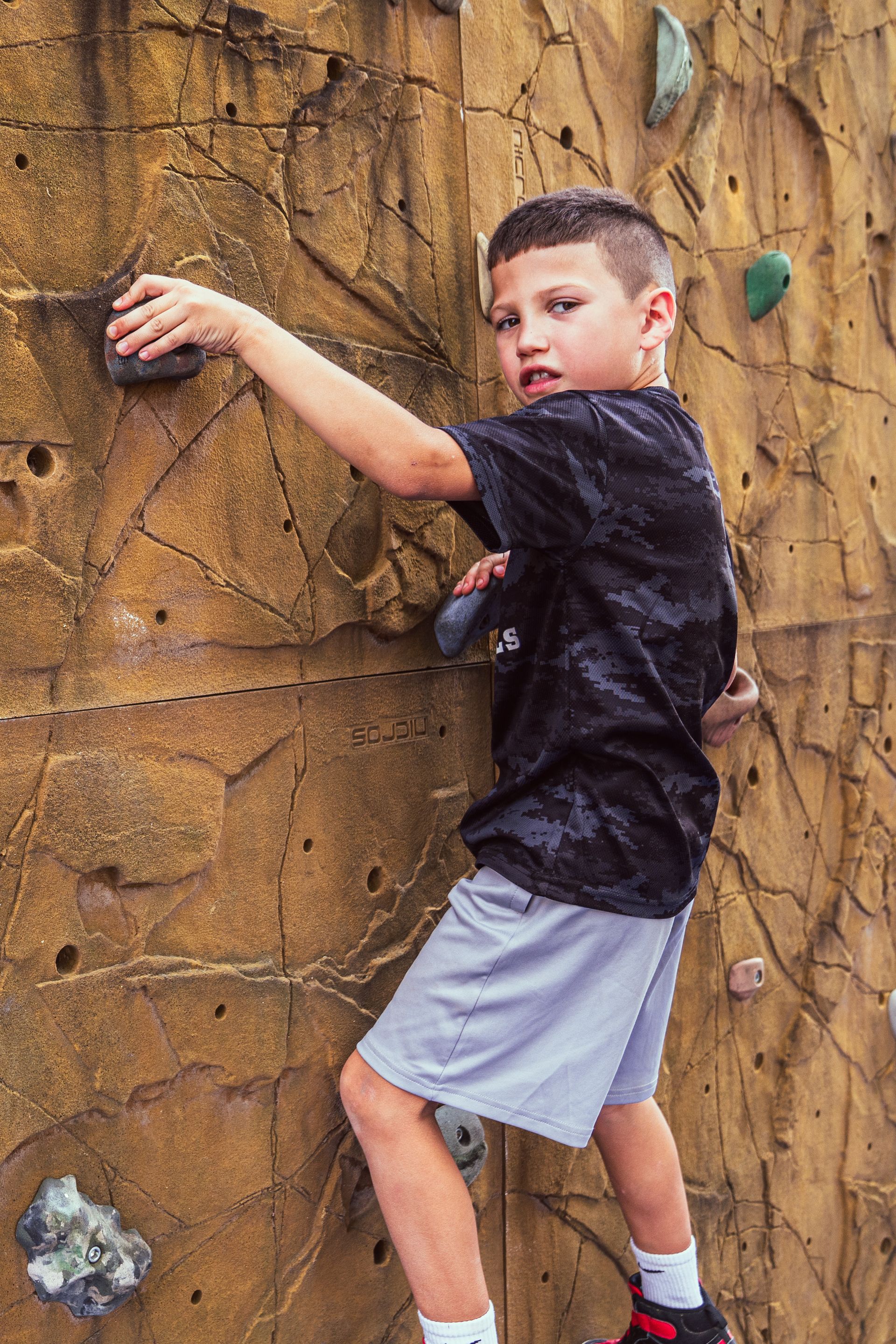 Boy climbing a beige rock climbing wall, reaching for a hold. He's wearing a black t-shirt and gray shorts, looking focused.
