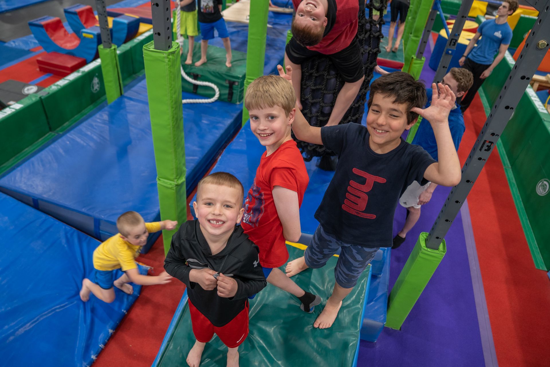 Children playing on a colorful indoor obstacle course. Boys wave and smile, while others play on the equipment.