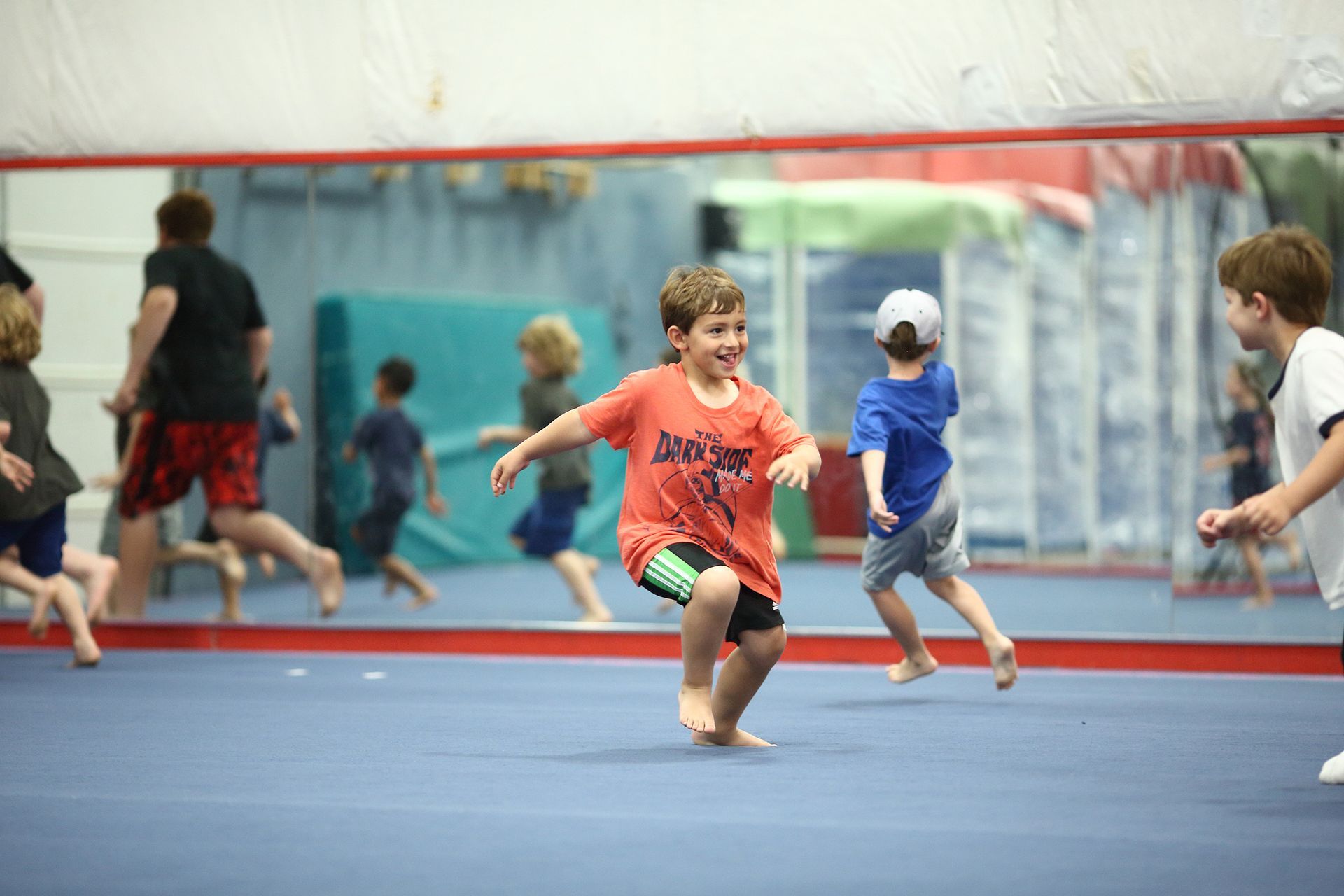 Children run and play on a blue gym floor, reflected in a large mirror. One boy in a red shirt is smiling.