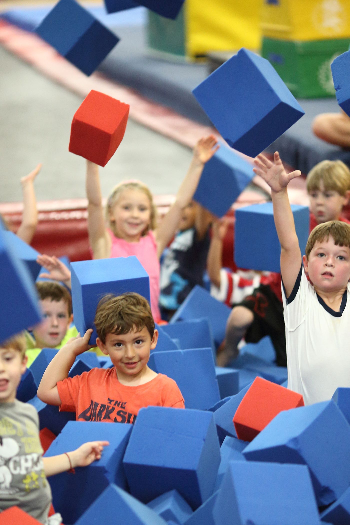 A young boy hangs from gymnastic rings indoors, looking up with a focused expression. The setting appears to be a fitness center with 