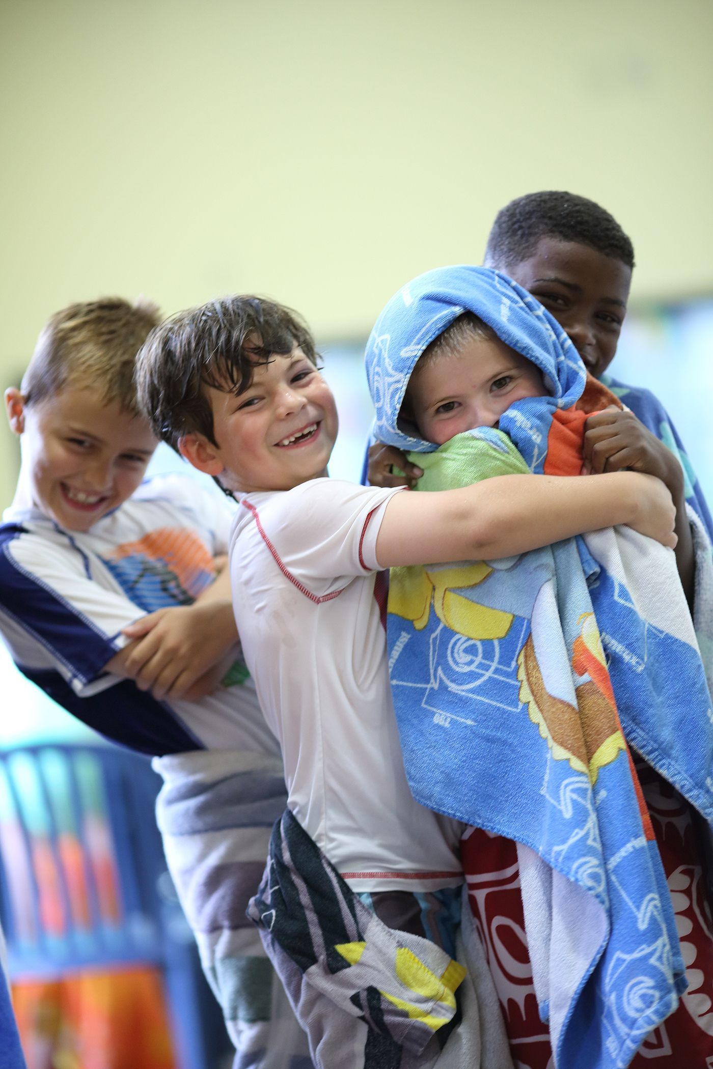 Four children, smiling, huddled together, one wrapped in a blue towel with a hood. Indoor setting.