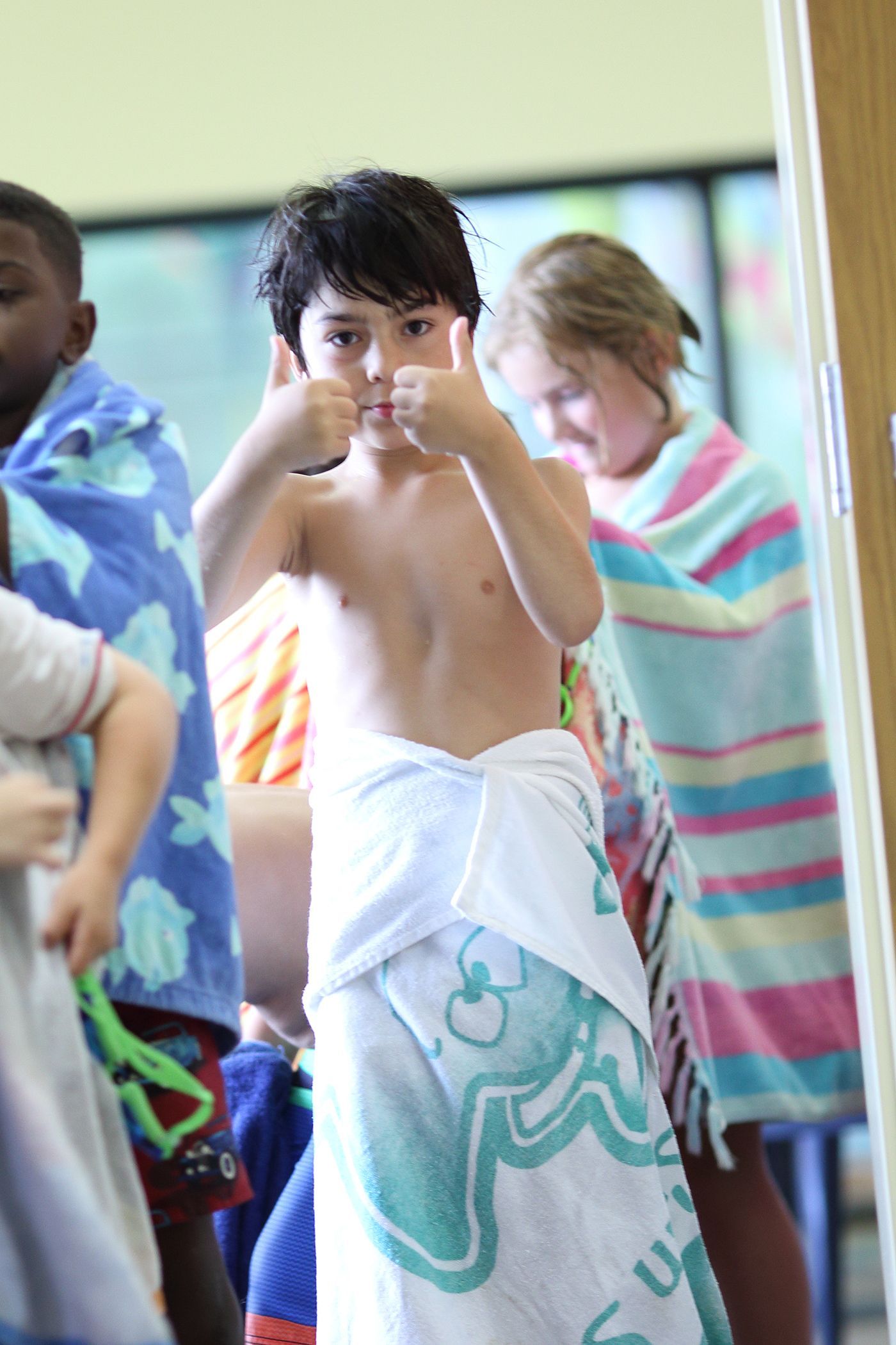 Boy in white towel gives thumbs-up after swimming; other children wrapped in towels are blurred in the background.