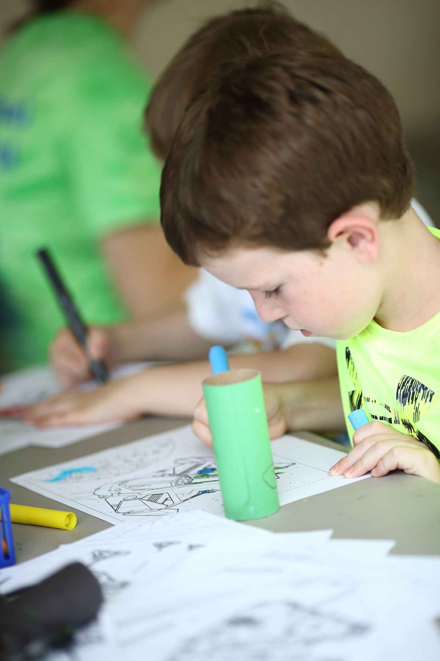 Boy with brown hair colors a picture at a table, holding a blue marker. Another person colors in the background.