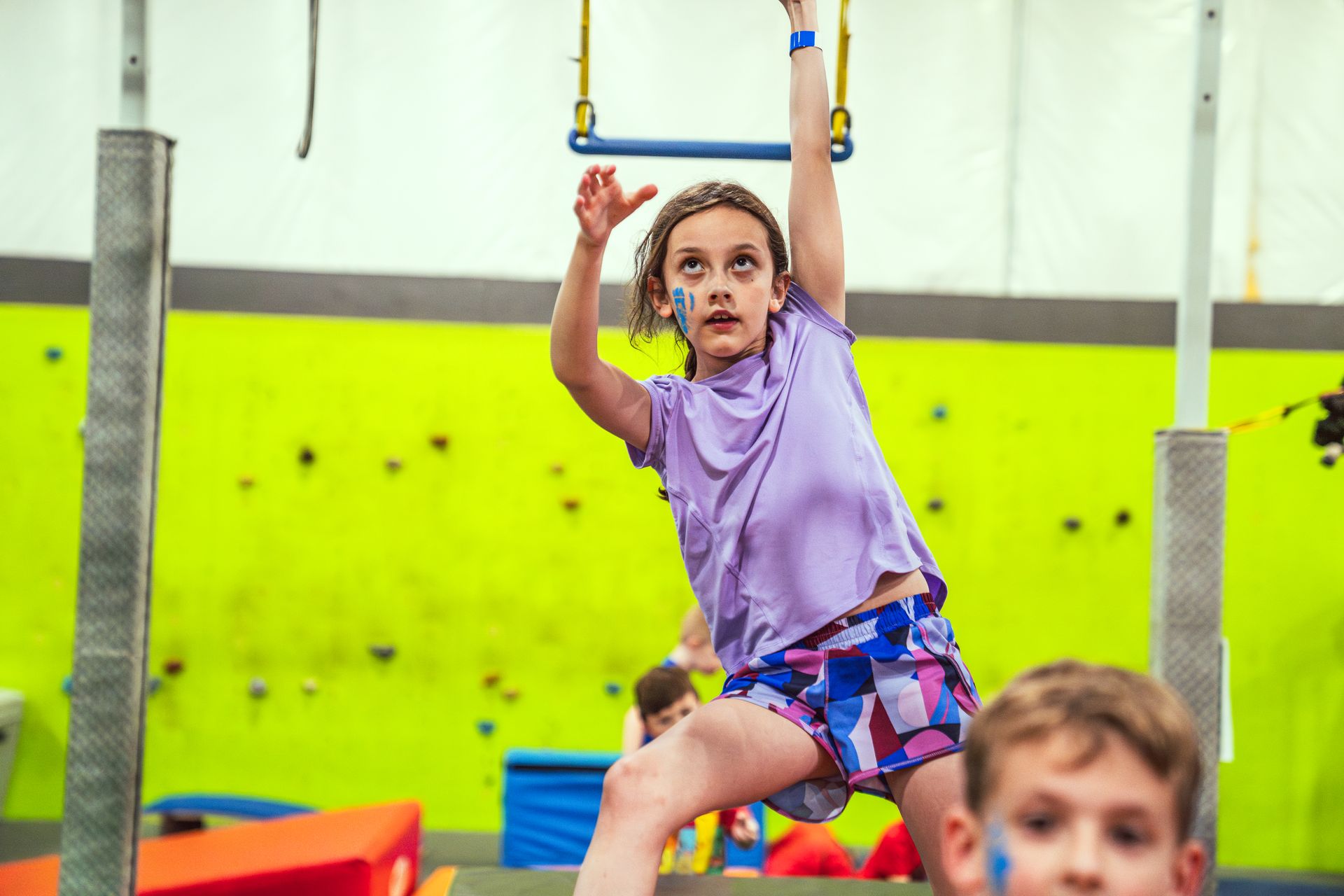 Girl in a purple shirt and shorts on a small trapeze indoors, with blue face paint.