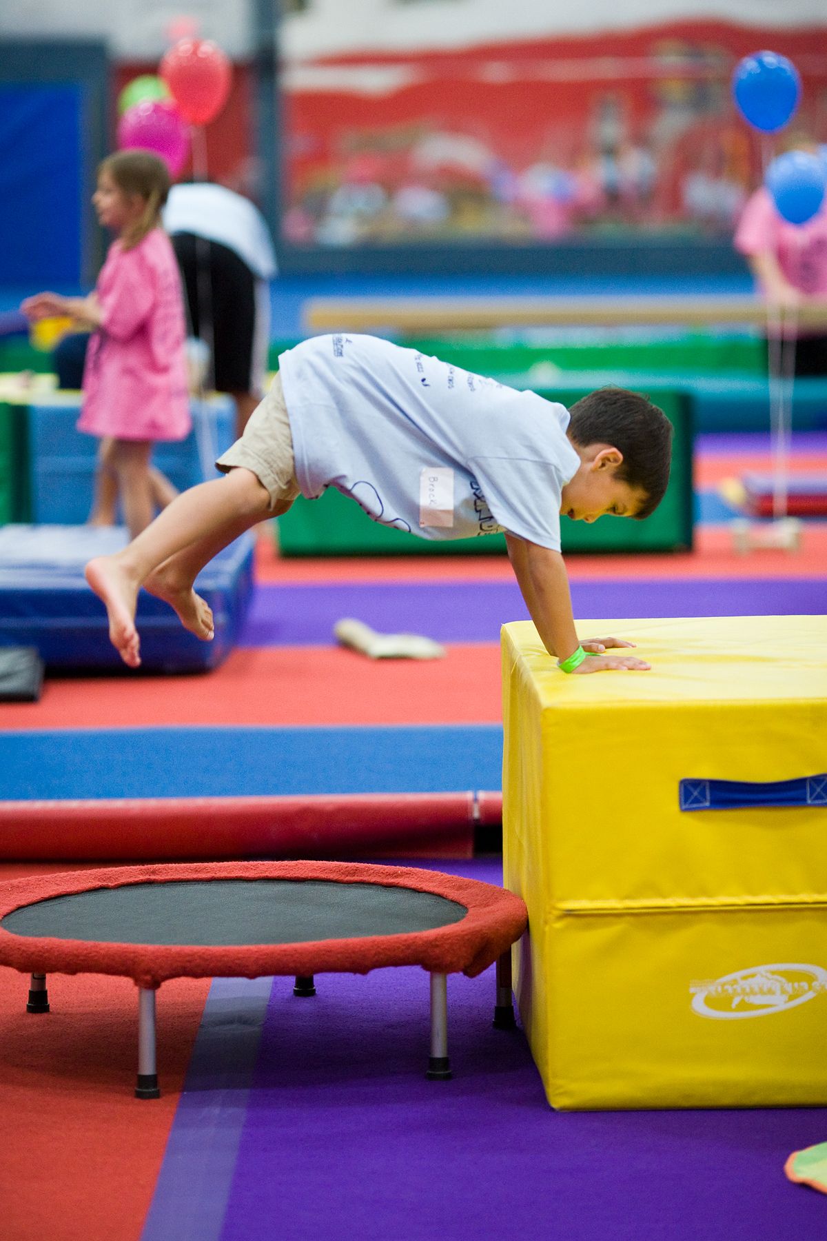 Boy doing a handstand on a yellow block, with a trampoline below. Other children in a gymnasium.