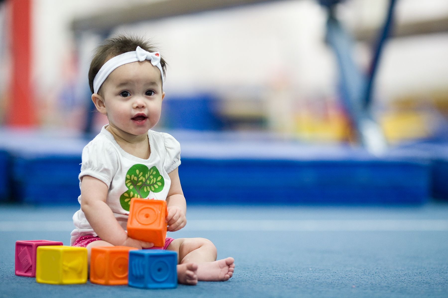 Baby sitting with colorful blocks on blue mat in a gym, looking towards the camera.