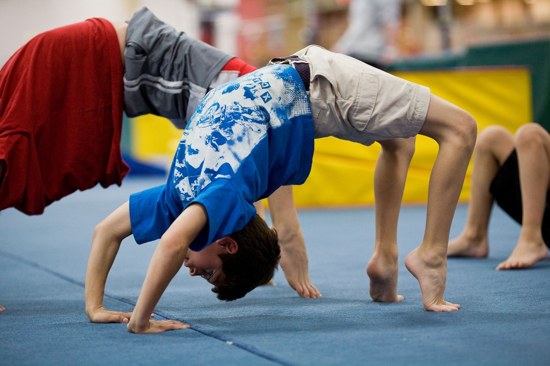 Boy in blue shirt doing a bridge on a blue mat, with a boy to his right and hanging clothing above.