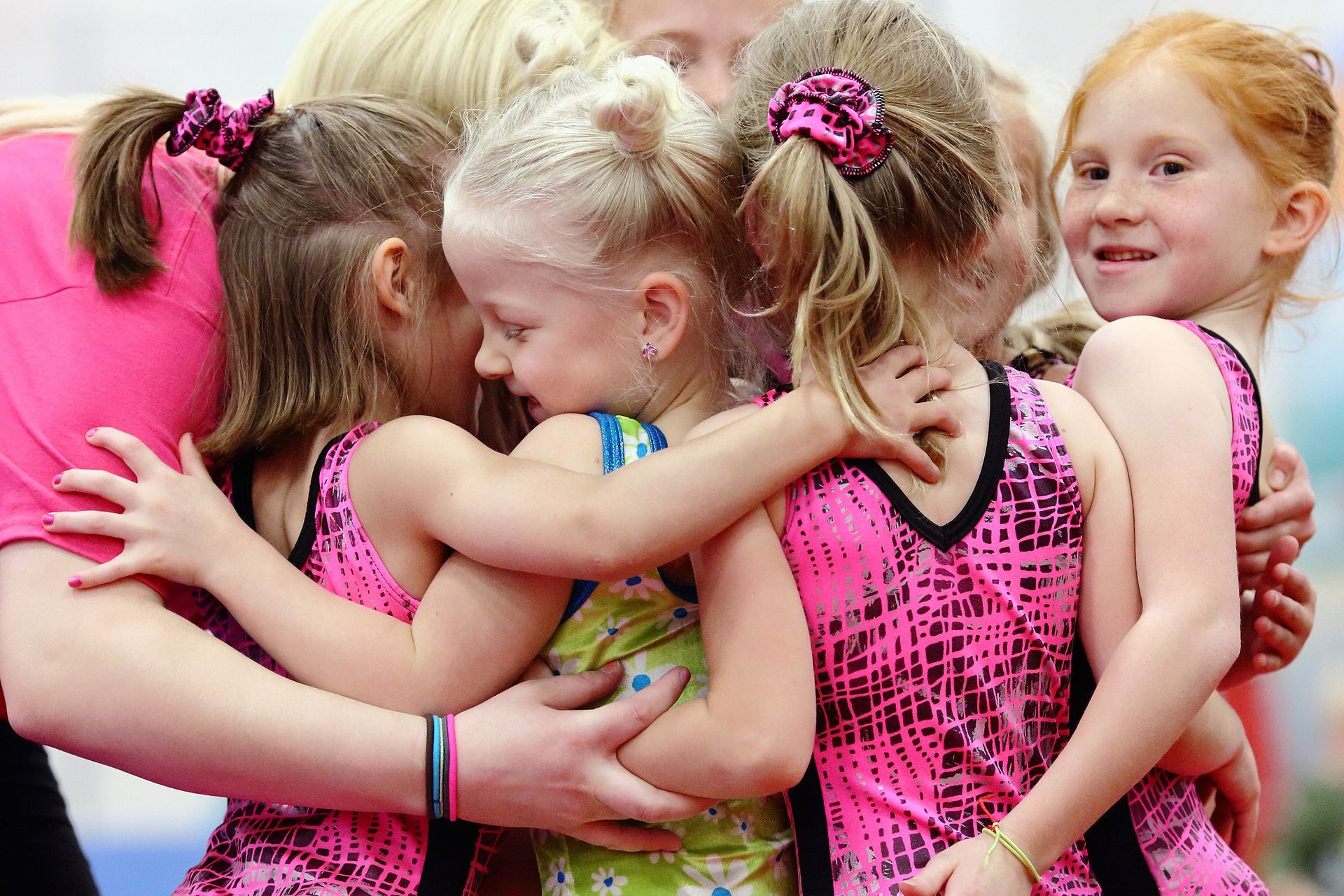 Young gymnasts in pink and black uniforms huddle together, smiling, with a coach.