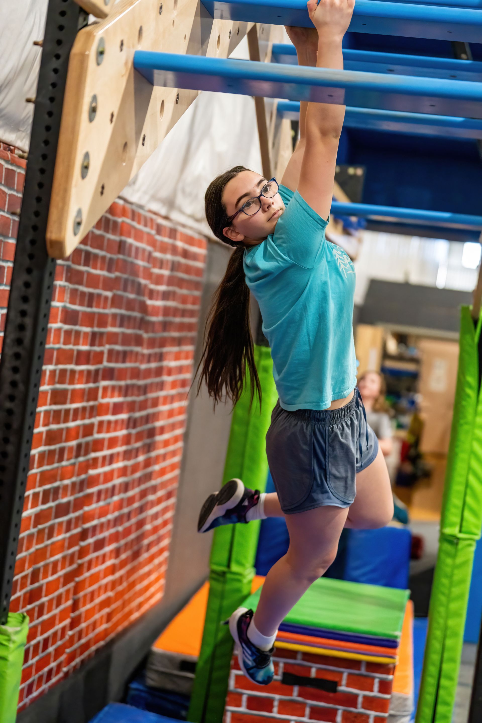 Girl with dark hair and glasses hanging from a blue bar on a climbing structure. She is wearing blue shorts and a teal shirt.