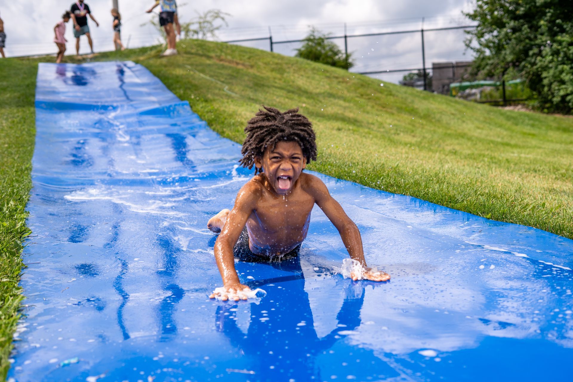 Young Black boy slides down a wet, blue slip-n-slide on a grassy hill, with an excited expression.