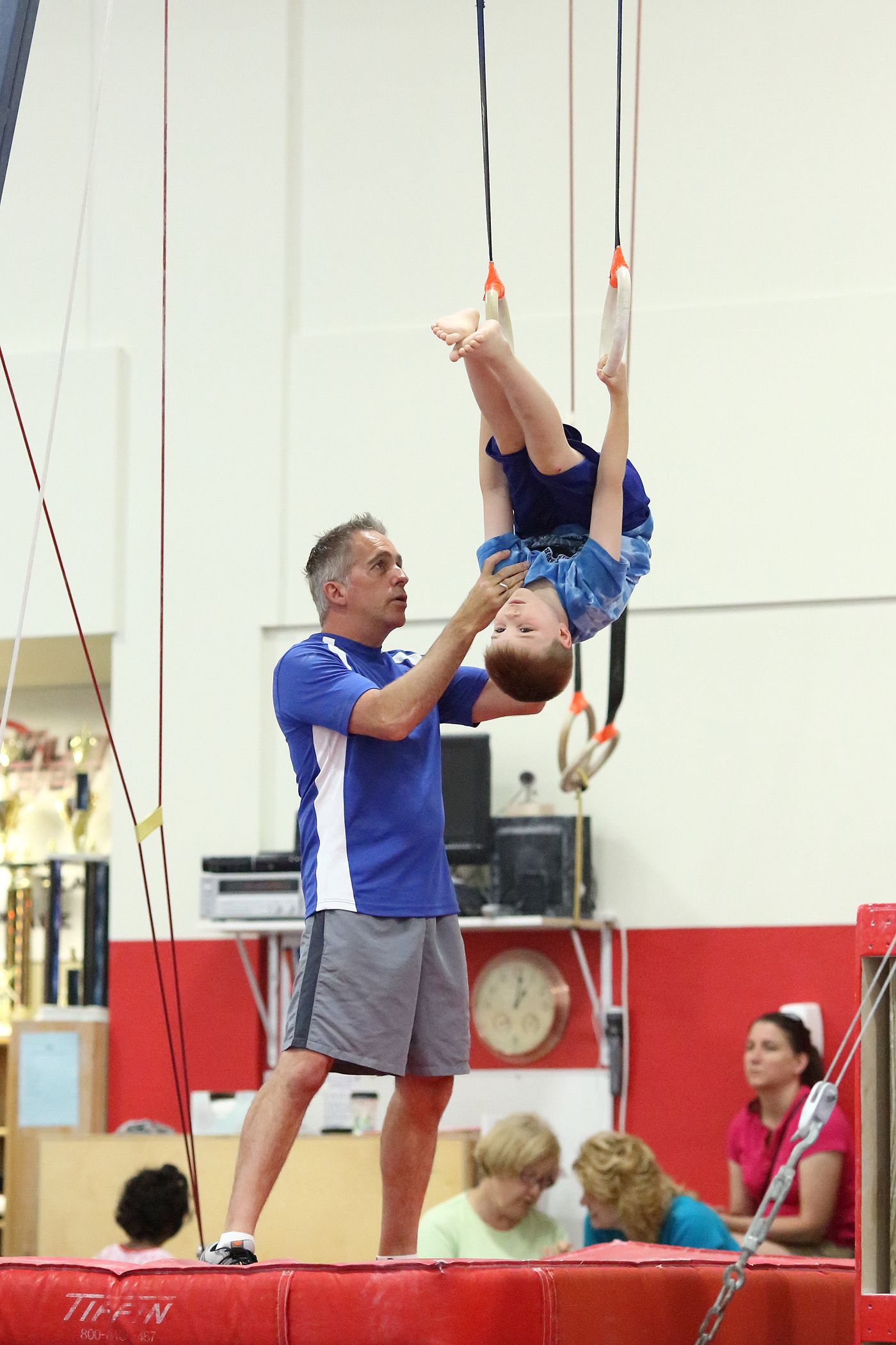 Gymnast on rings with a coach, red mat and audience in background.