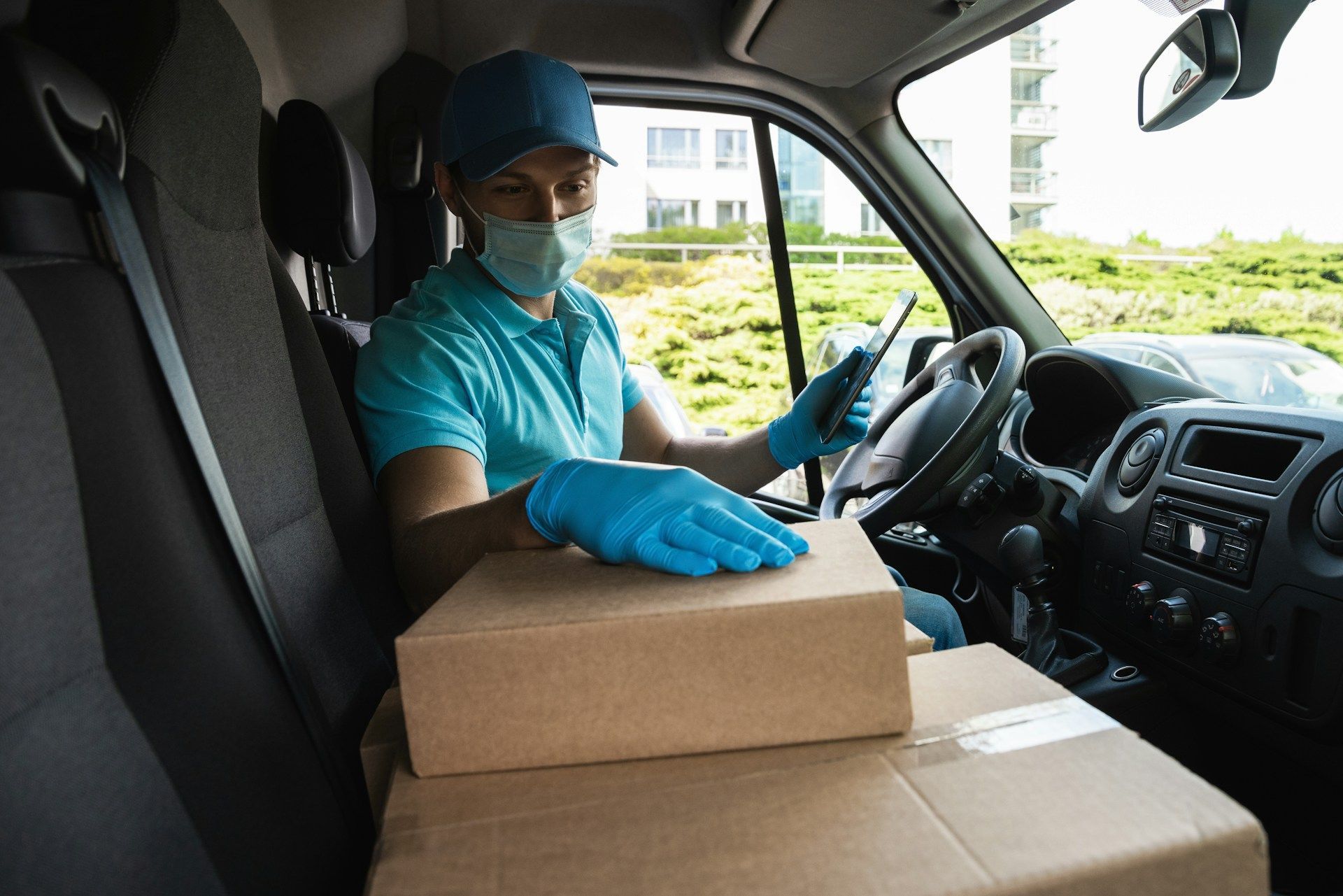 A delivery man wearing a mask and gloves is sitting in a van with boxes.