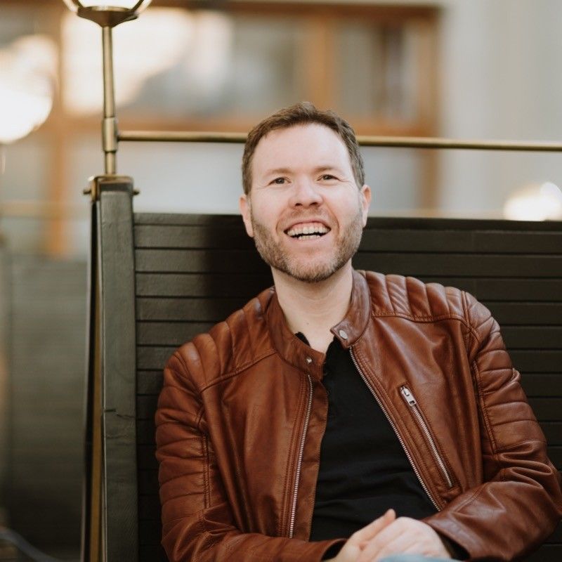 A man in a brown leather jacket is smiling while sitting in a chair