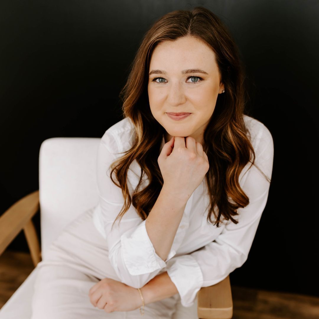 A woman in a white shirt is sitting in a chair with her hand on her chin.
