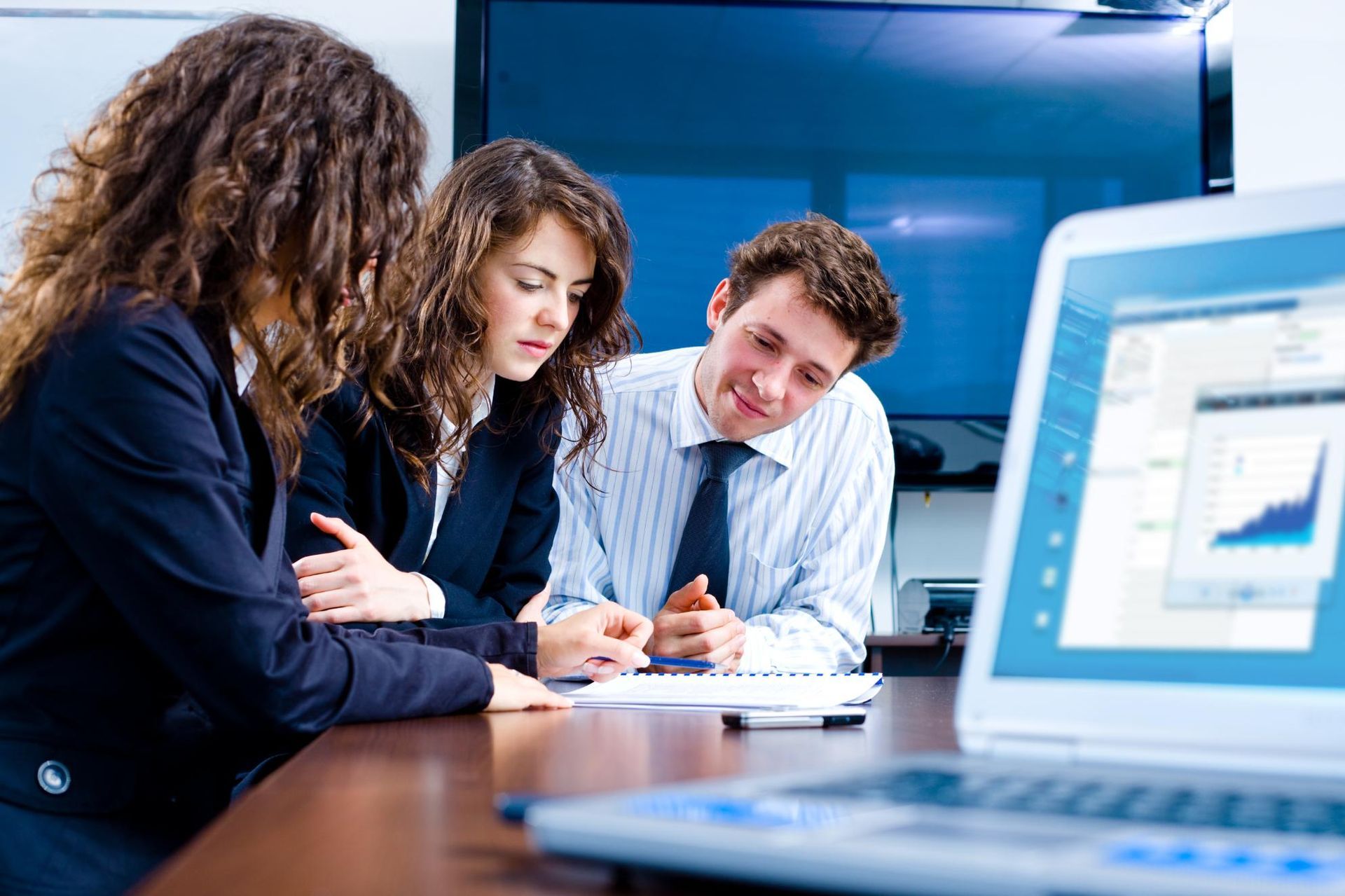 Three businesspeople in a meeting, reviewing documents and looking at a laptop screen displaying a graph.