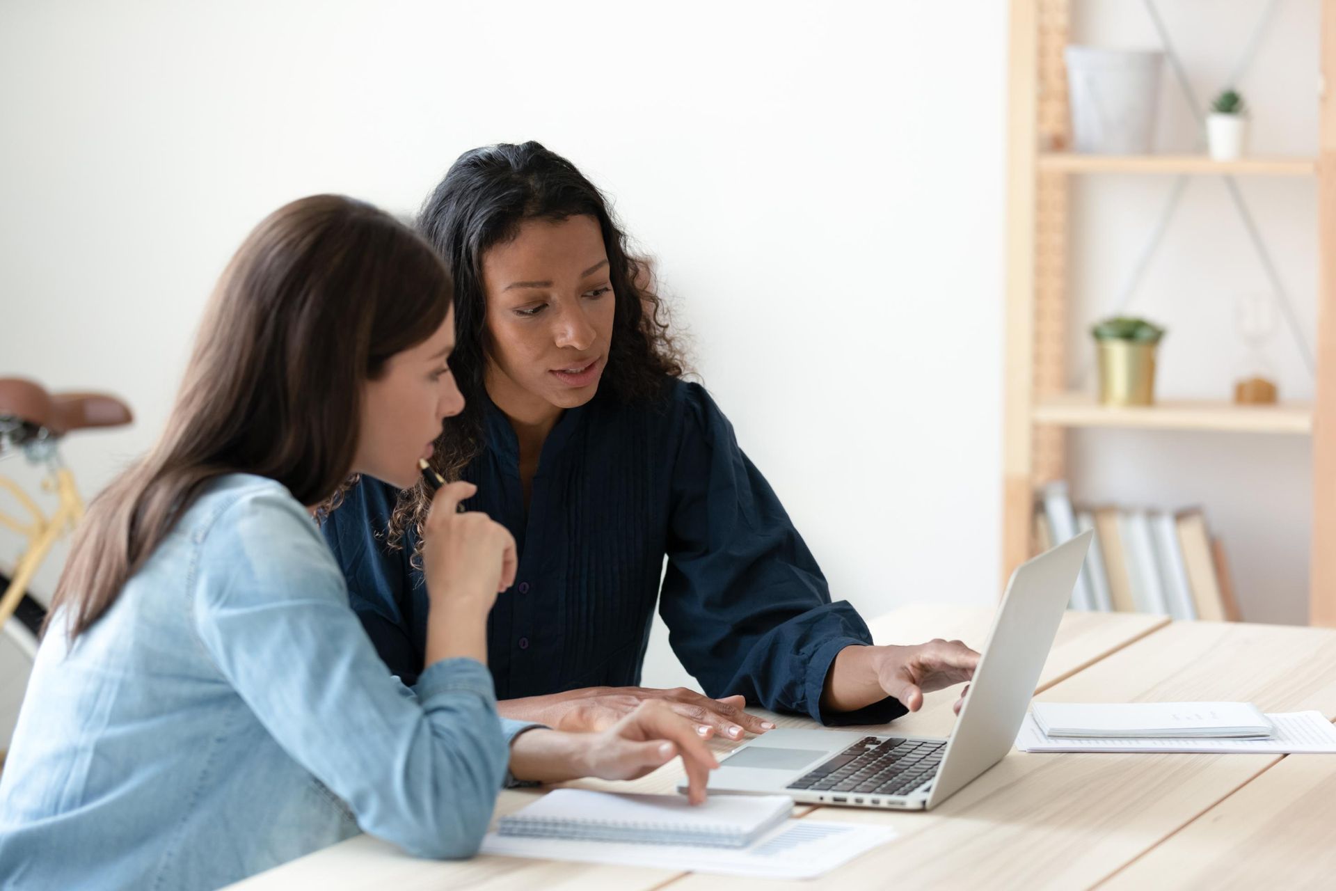 Two women reviewing data on a laptop at a wooden table, with one pointing.