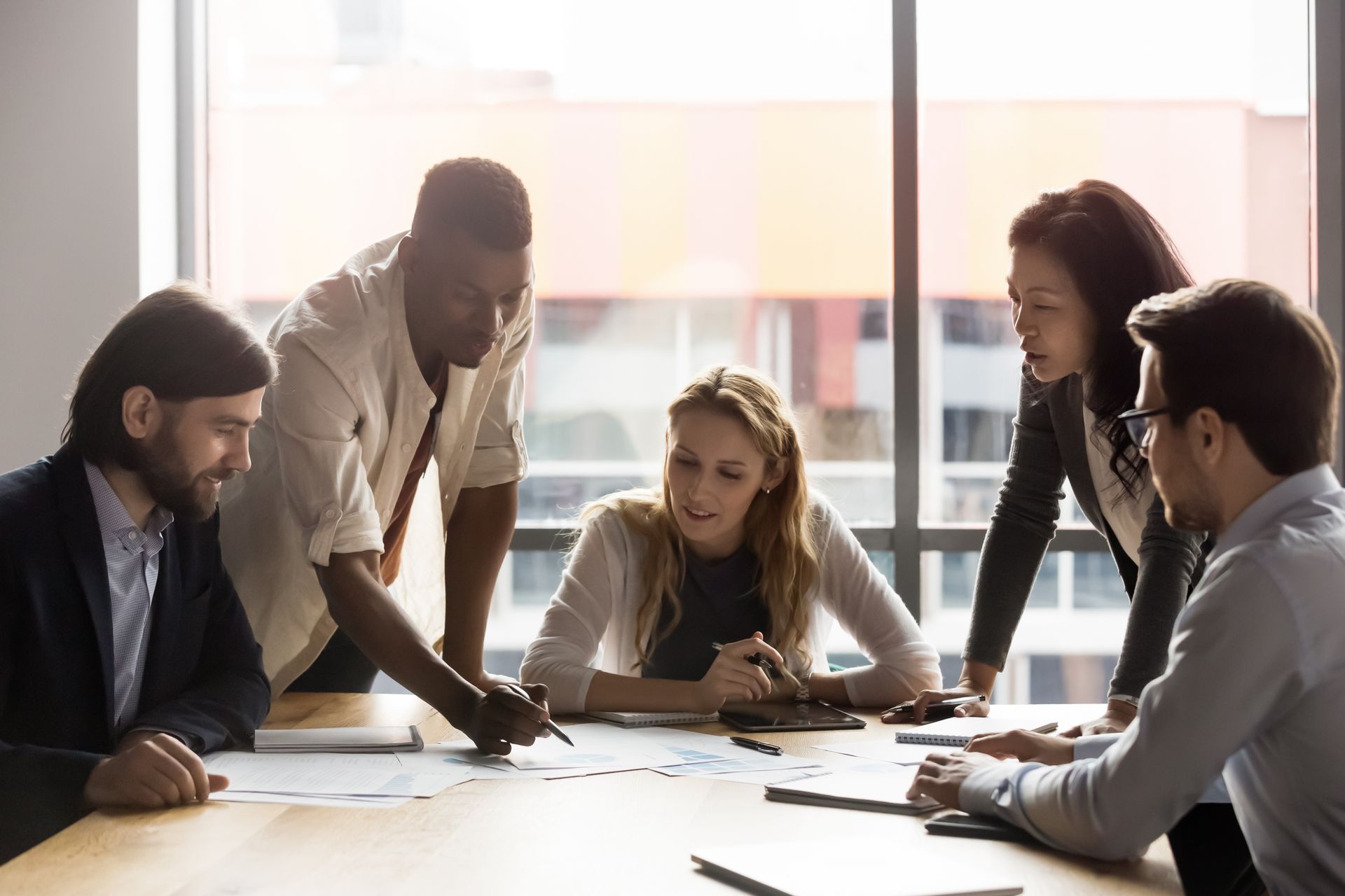 A diverse team reviews paperwork around a table in a sunny office. They point, analyze, and collaborate.