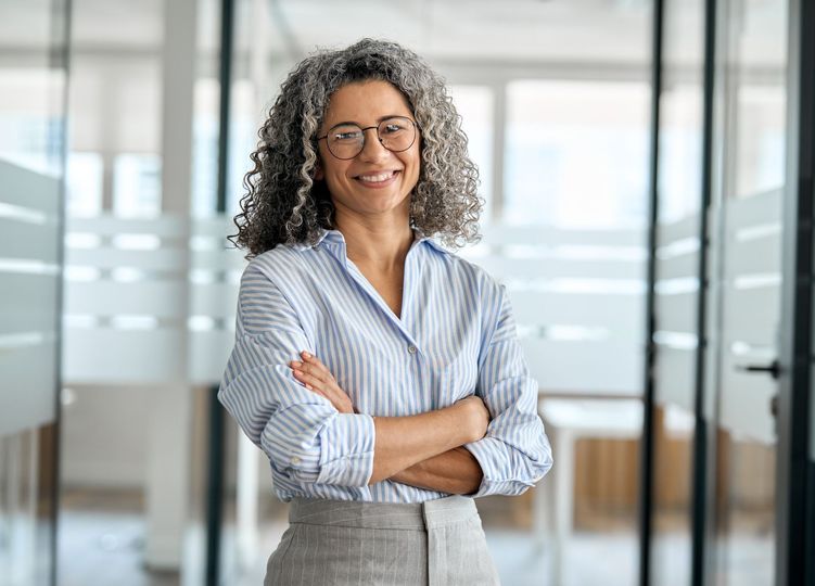 Woman with gray curly hair and glasses smiles, arms crossed, in a modern office hallway.