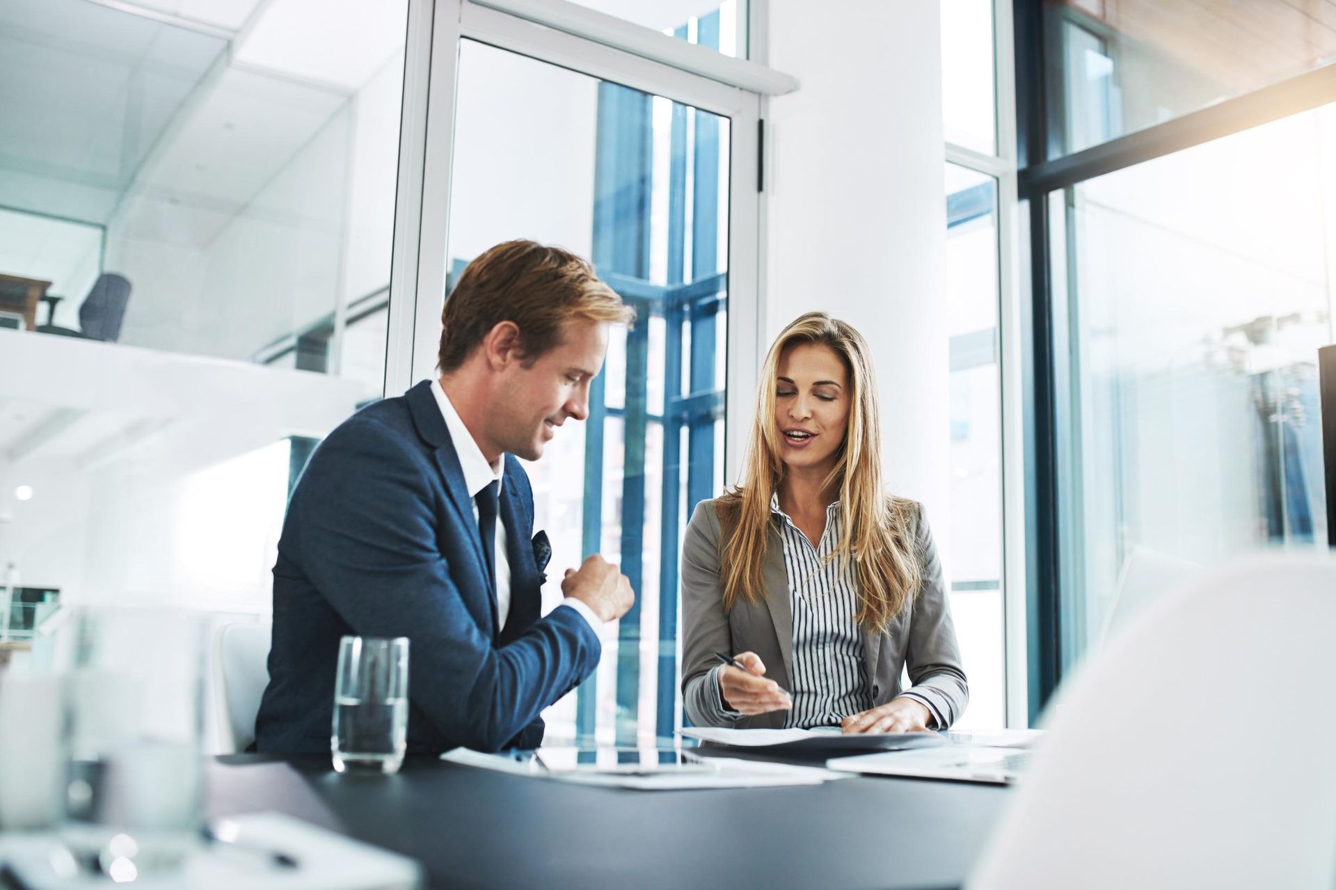 Man and woman in business attire reviewing documents at a table in a modern office setting.