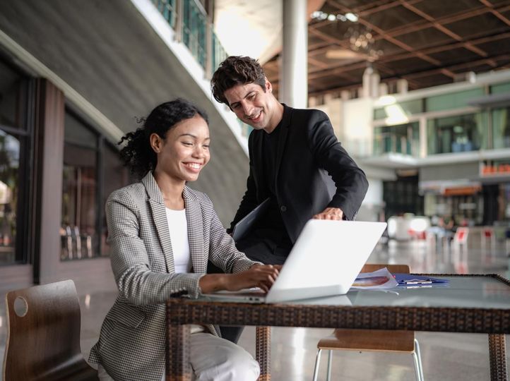 A woman and a man smile while looking at a laptop in a light-filled building. 