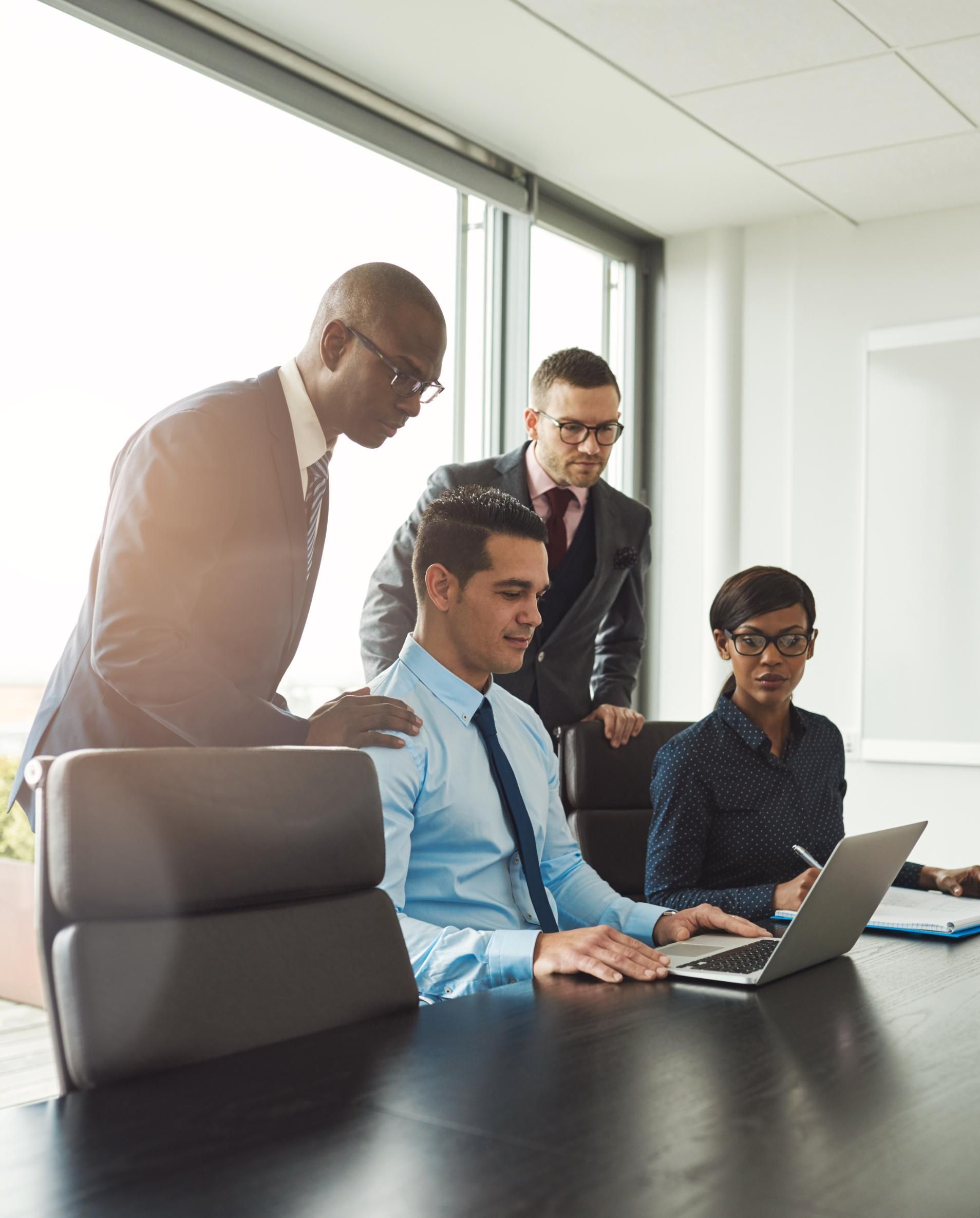 Four business people in suits gather around a laptop in a modern office. 