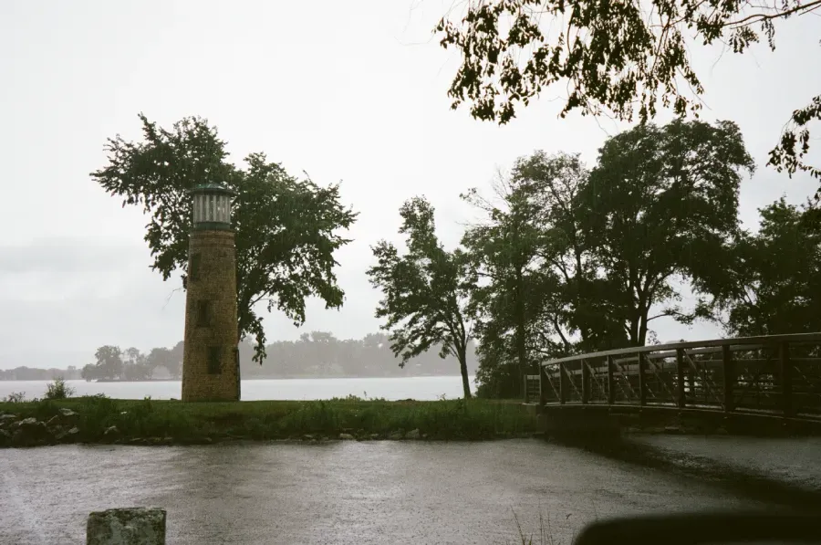 Asylum Point Dink stands on a grassy shore by a lake, with trees and a wooden footbridge under an overcast, rainy sky.