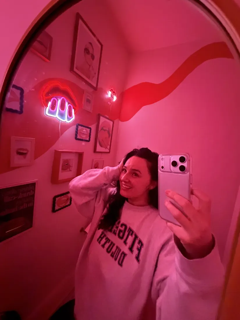 Person taking a mirror selfie in a red-lit room with framed wall art and a neon sign.