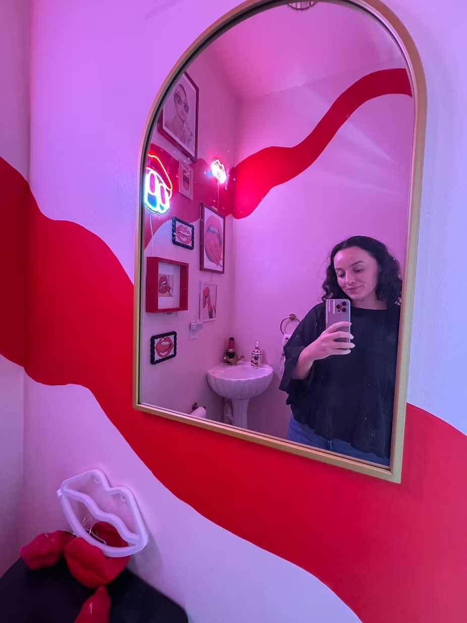 Charlee Remitz taking a selfie in a red-lit bathroom with red wave-like patterns. Gold-framed mirror
