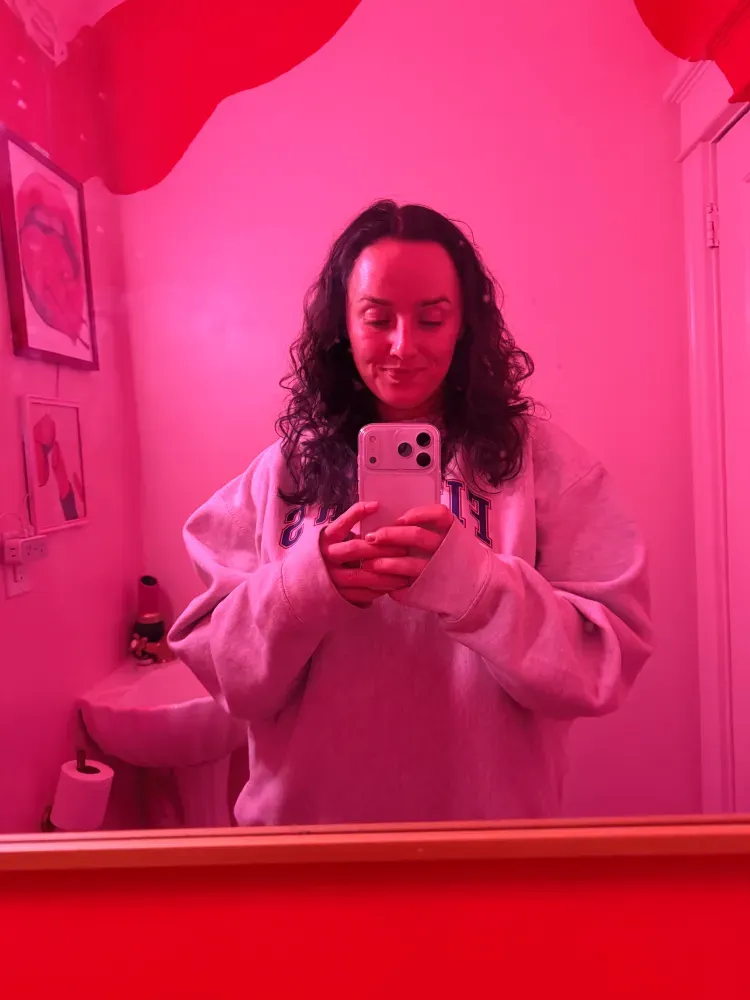 Charlee Remitz wearing a sweatshirt takes a mirror selfie in a room lit with pink light.