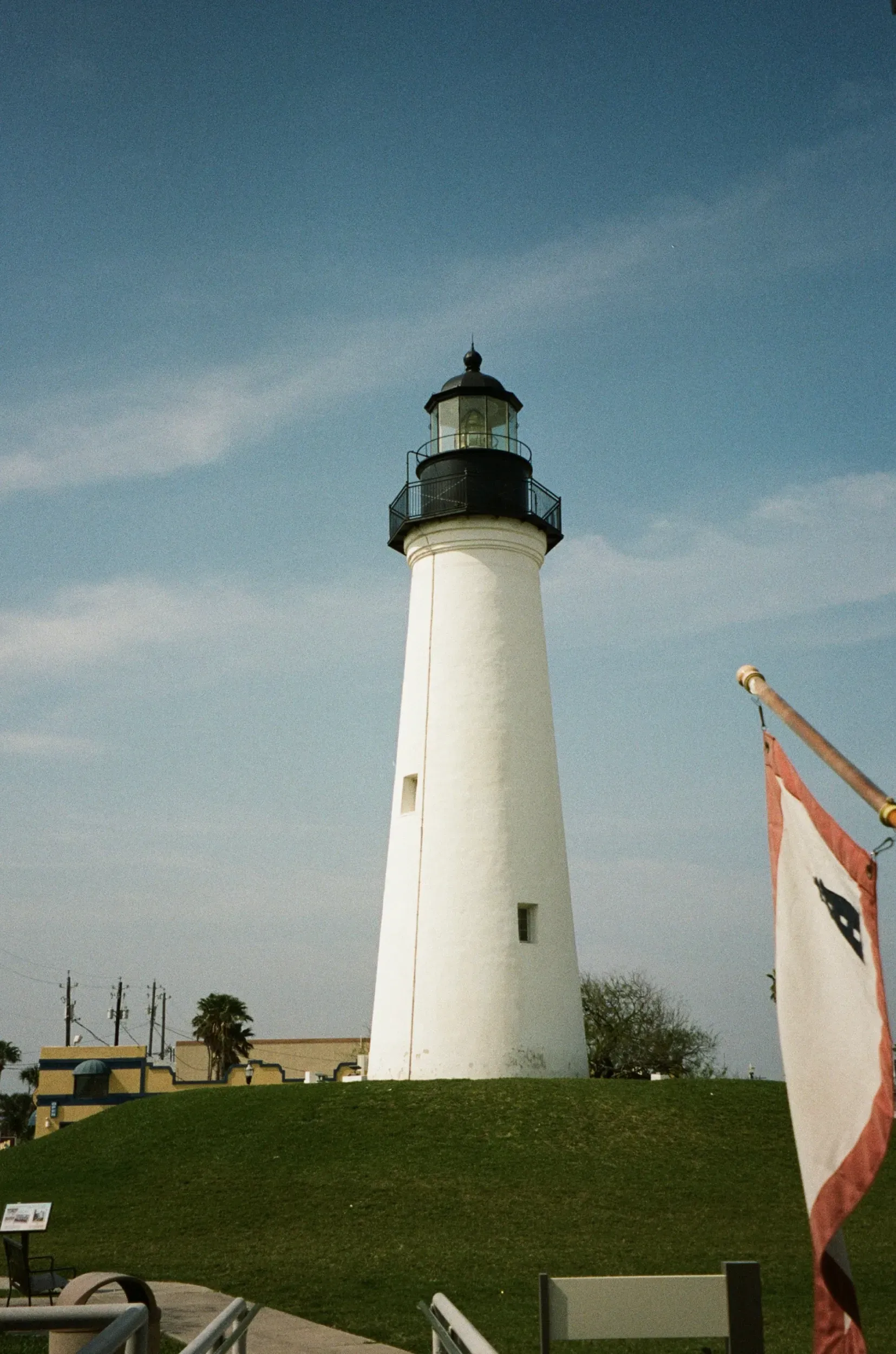 White lighthouse on a grassy hill under a blue sky, with a flag and distant buildings.