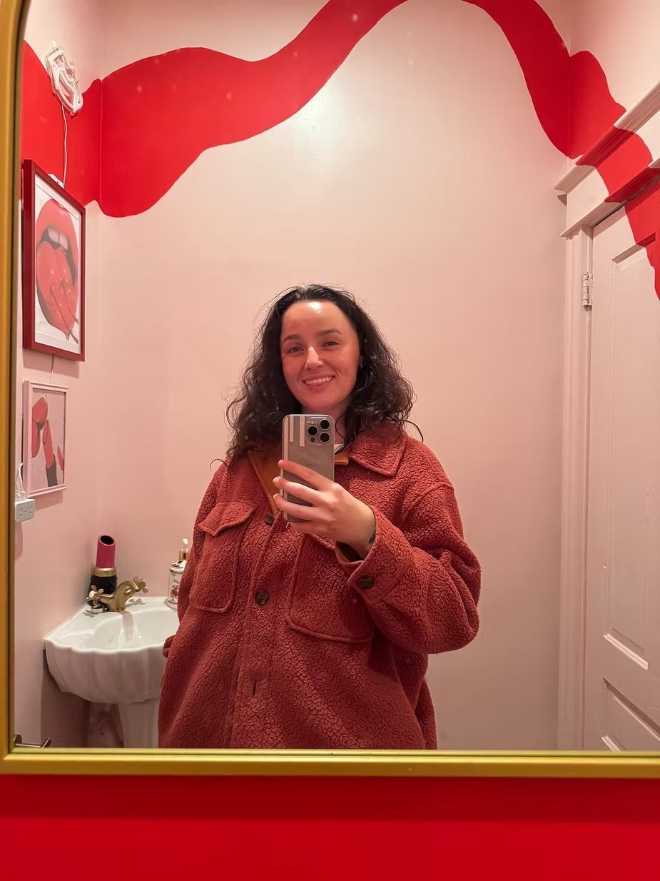Woman in red jacket taking a selfie in a pink-themed bathroom with red lip art on the wall.