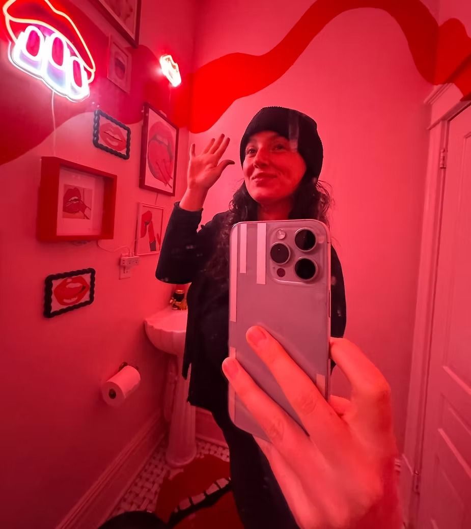 Charlee Remitz in black hat takes selfie in red-lit bathroom, waving.