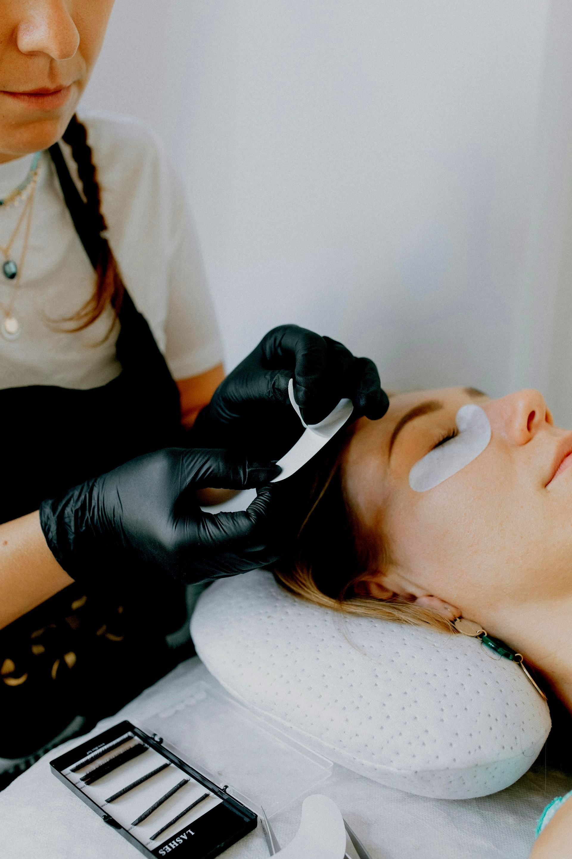 A woman is getting her eyelashes done by a woman wearing black gloves.