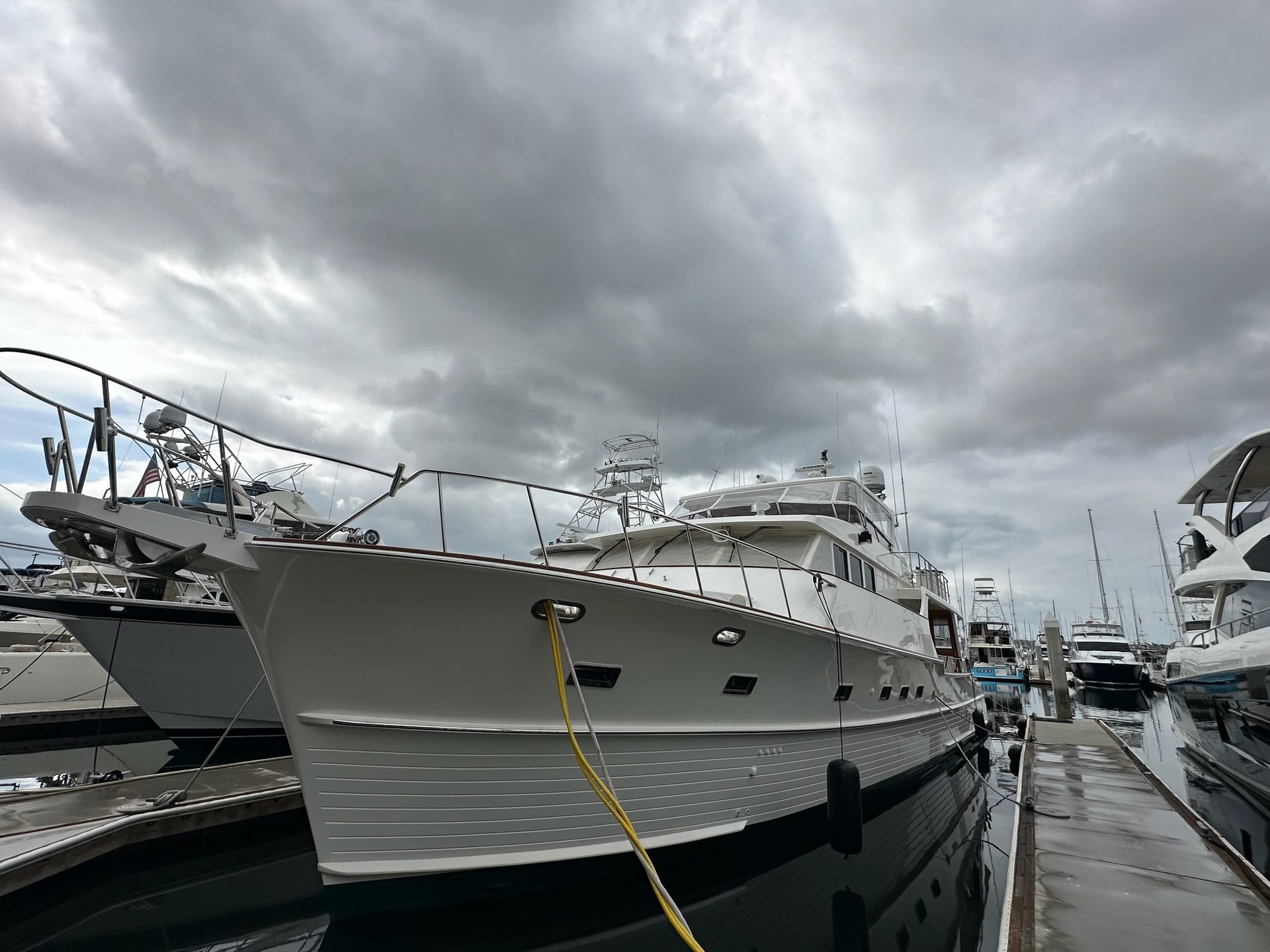 White yacht docked in a marina under an overcast sky. The boat is secured with yellow ropes.