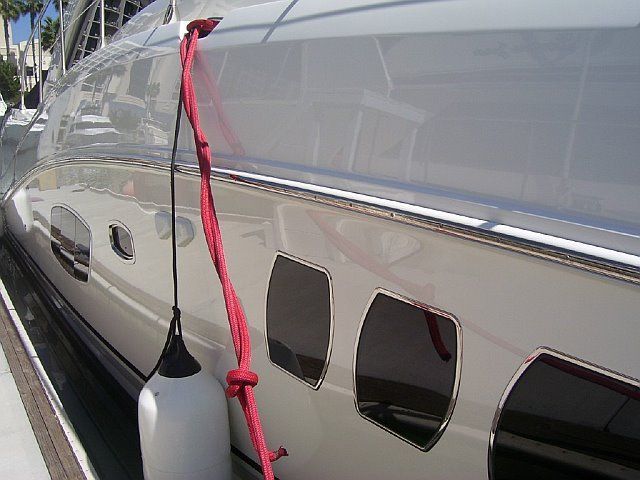 White yacht with a red rope and fender tied to it. The boat is docked, with several rectangular windows visible.