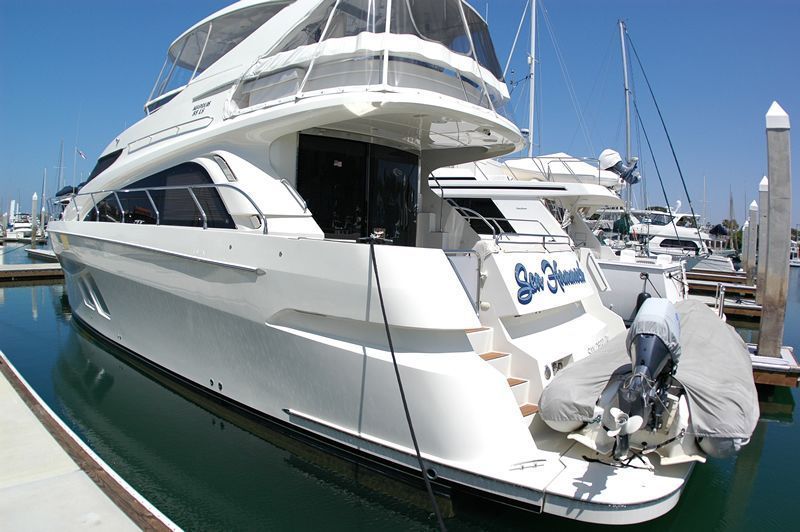 White yacht docked at a marina, stern facing the camera. Blue water, clear sky, and other boats are visible.