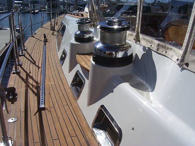 Close-up of a yacht's deck with teak wood and stainless steel winches. Sunlight reflects off the polished surfaces.