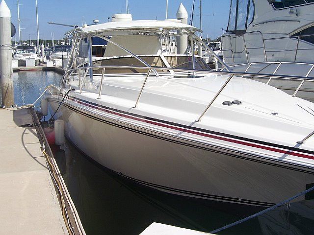 White motorboat docked in a marina, with a white canopy, red and black stripe, and other boats in the background.