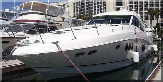 A white yacht docked in a marina with other boats and a tall building in the background.