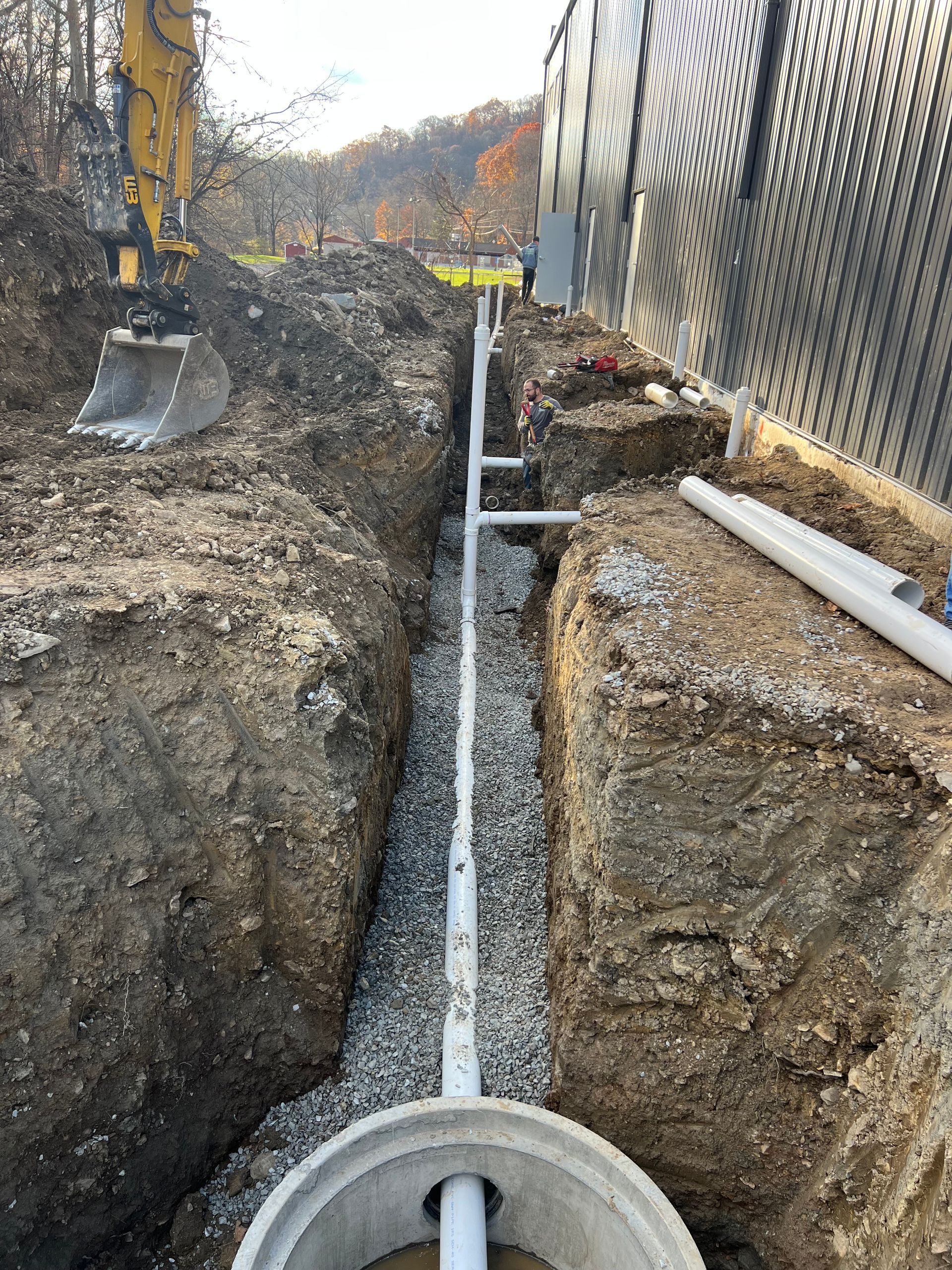 Trench with a drainage pipe system being installed next to a building. An excavator is in the background.