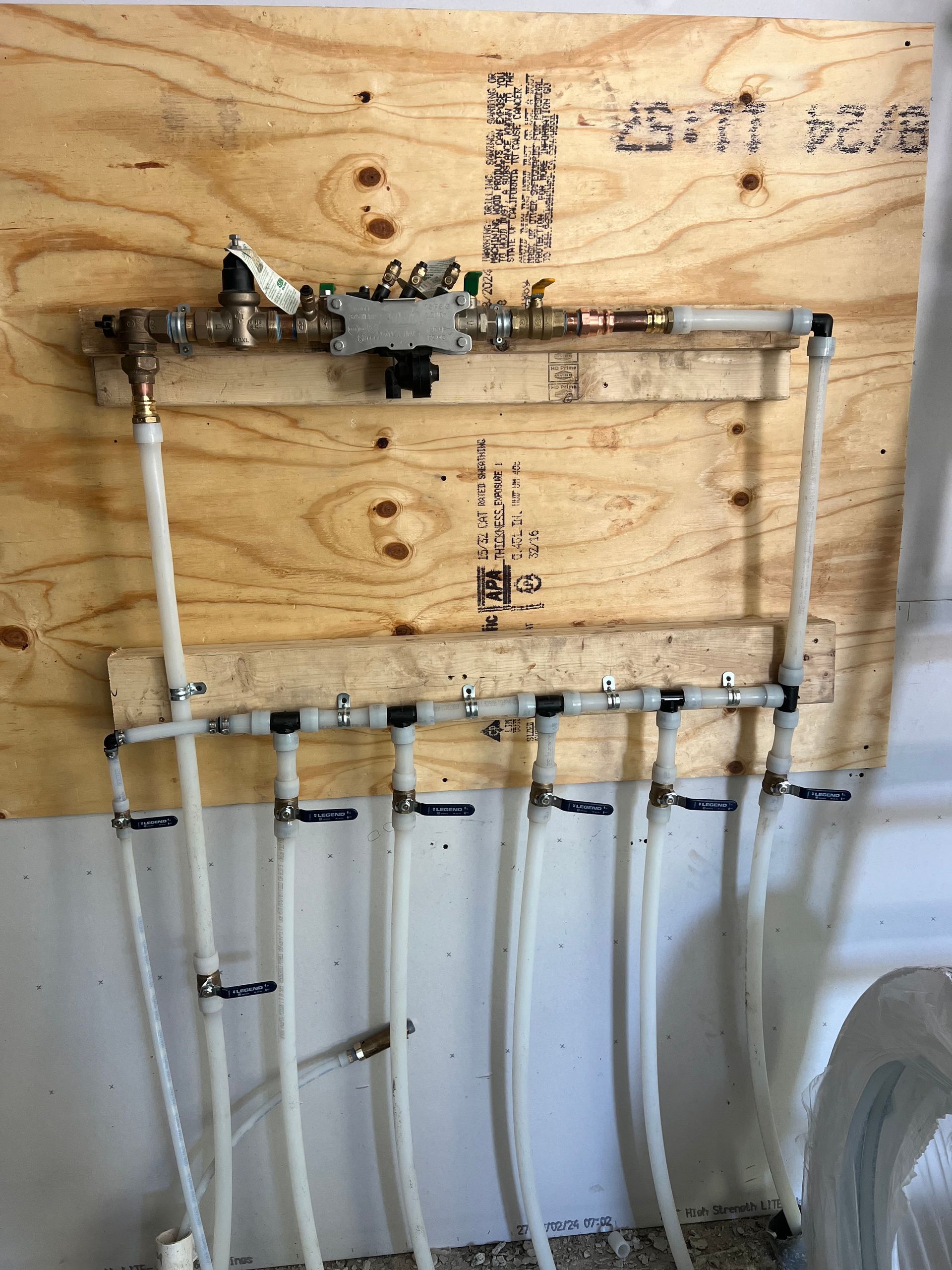 Radiant floor heating manifold system with white pipes and black valves mounted on plywood.