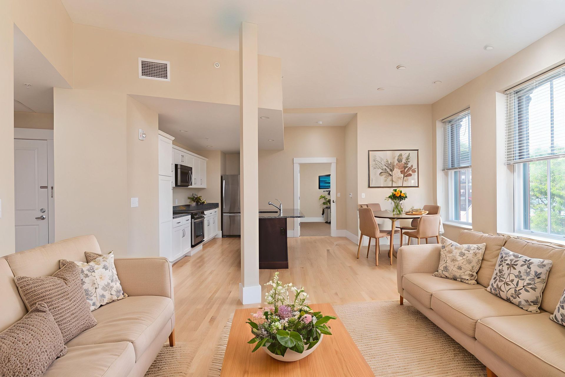 A modern, light-filled open-plan living room with two beige sofas, a wooden coffee table, and a kitchen in the background.