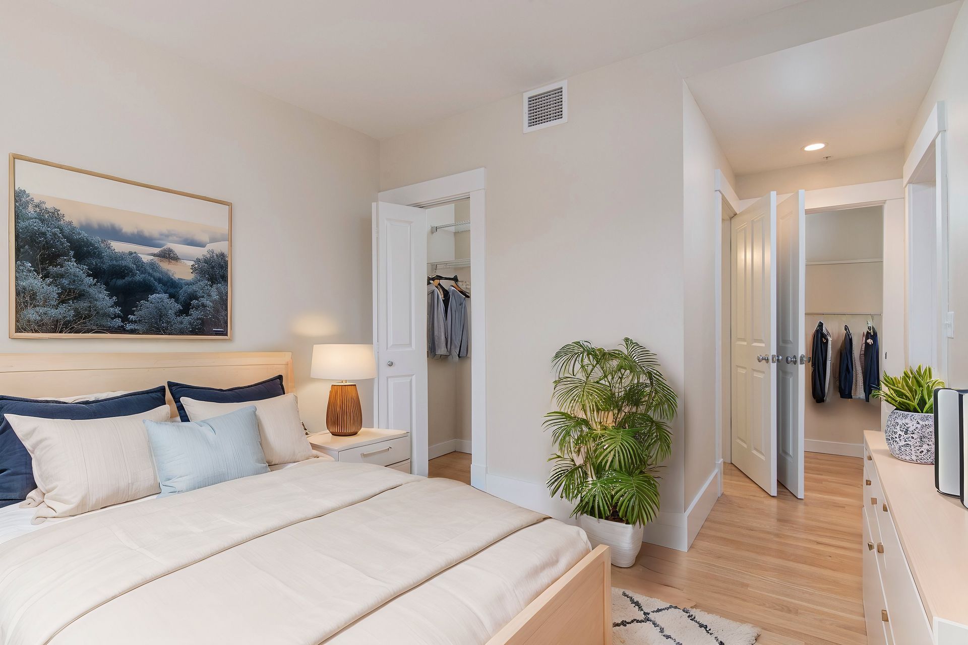 A bedroom with a queen bed, beige walls, a closet, a tall potted plant, and an open doorway to another room.