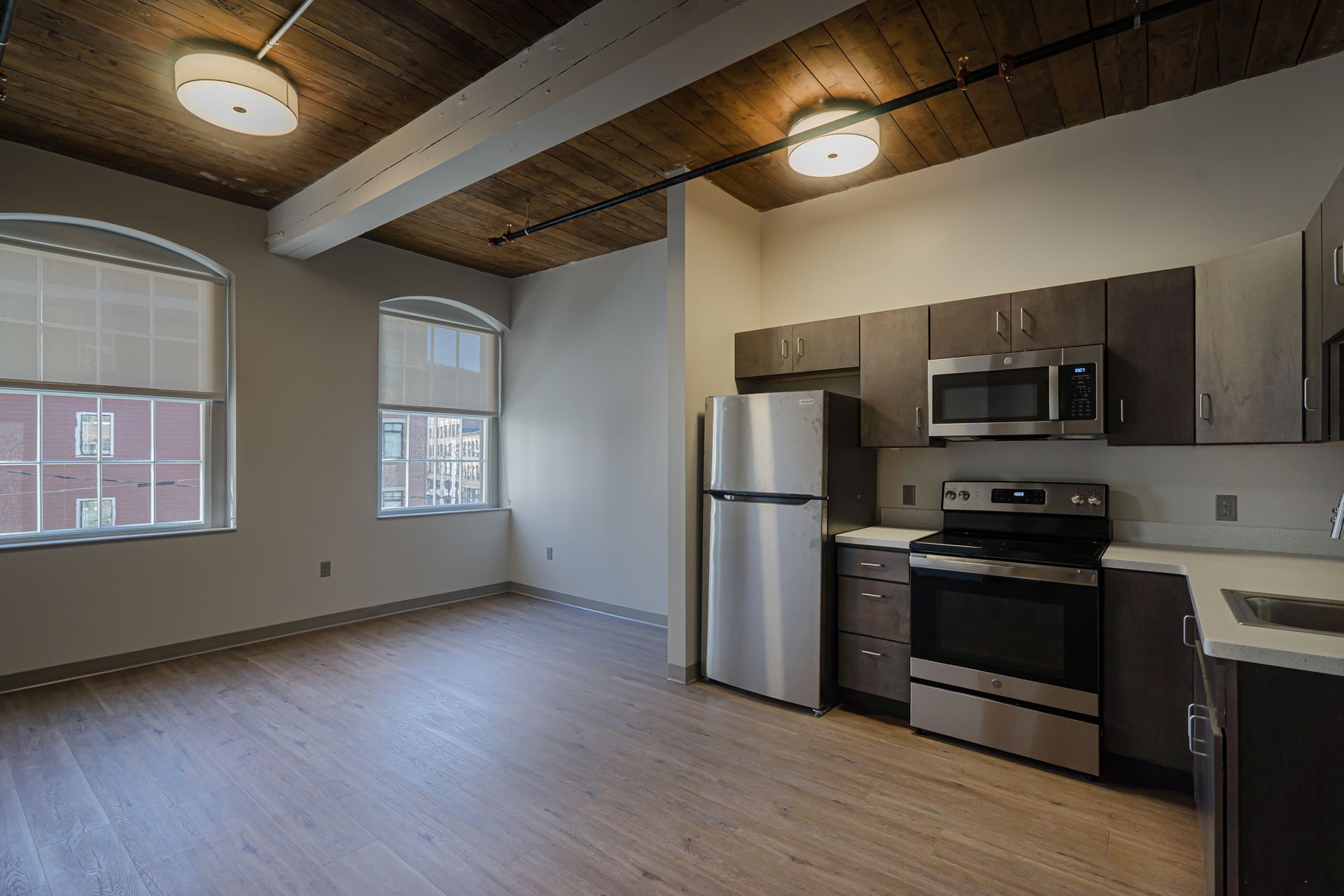 Empty apartment kitchen and living area with stainless appliances, wood floors, and exposed wood ceiling beams