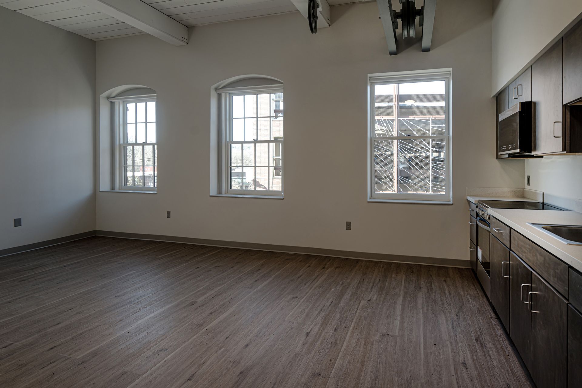 Empty apartment living room with wood floors, three windows, and a kitchen area on the right