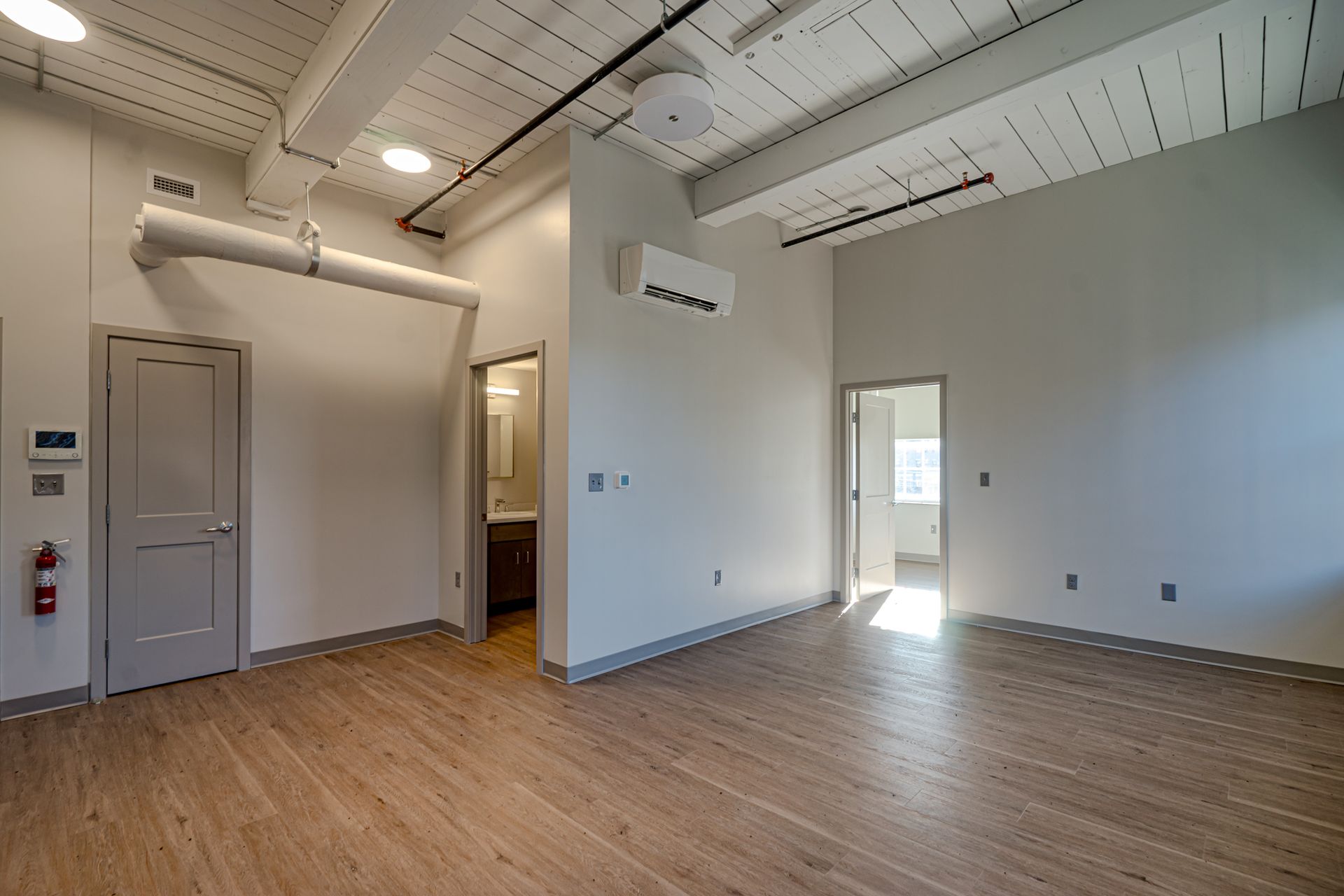 Empty loft-style room with wood floors, white walls, exposed ductwork, and daylight from a doorway.