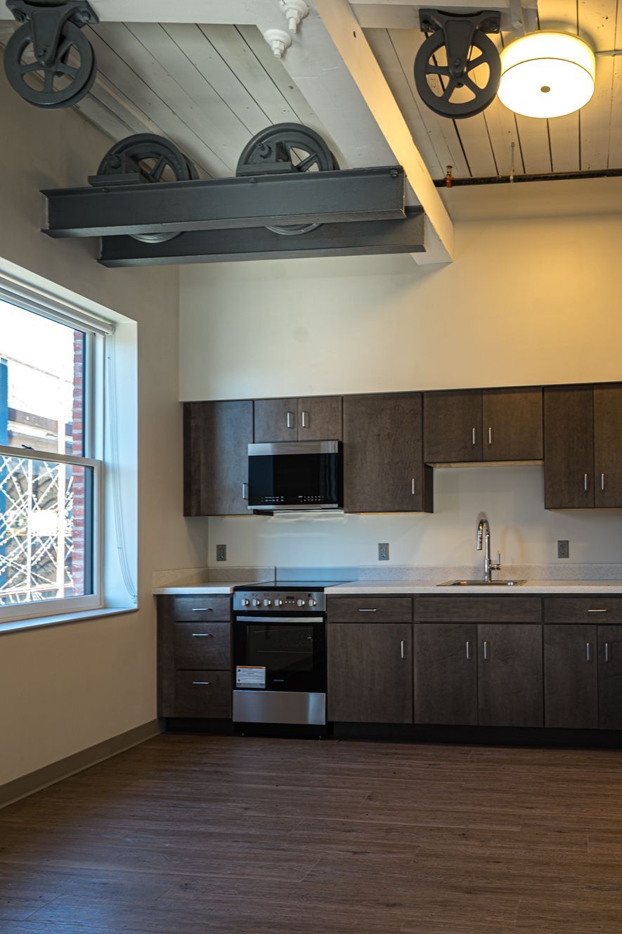 Small modern kitchen with dark cabinets, stainless stove, window, and decorative wagon wheels overhead