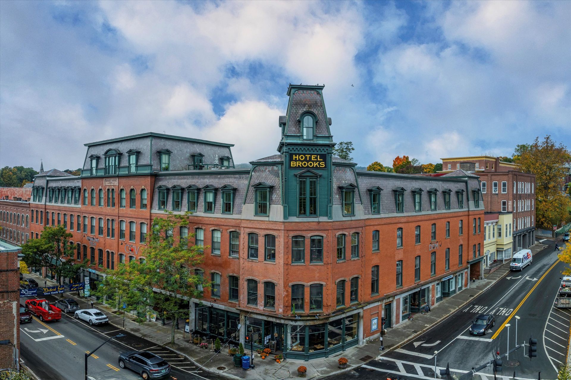 A four-story brick building with a mansard roof and a central clock tower, situated on a street corner under a blue sky.