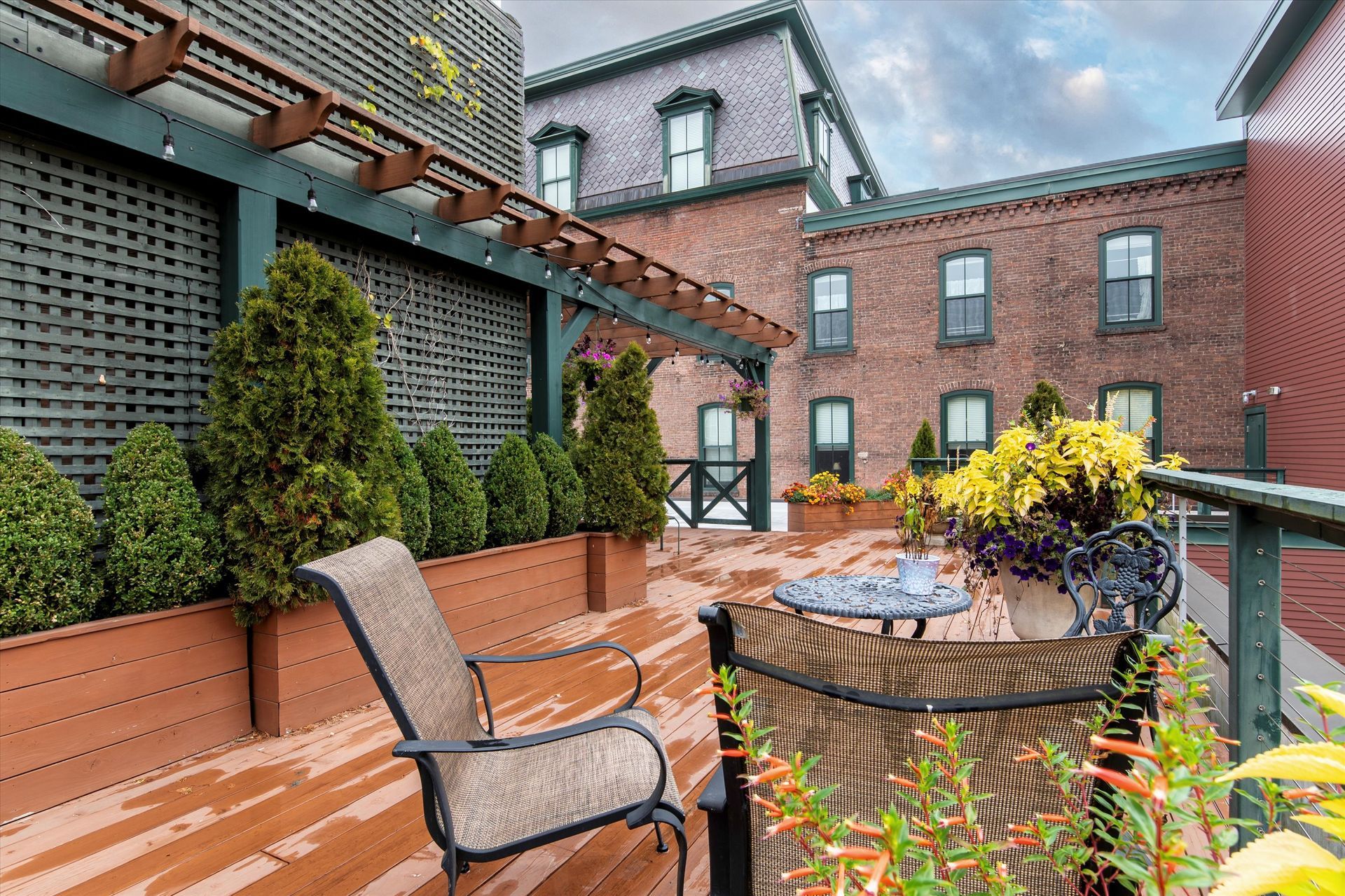 A patio deck with potted evergreen shrubs, outdoor seating, and a brick building in the background.