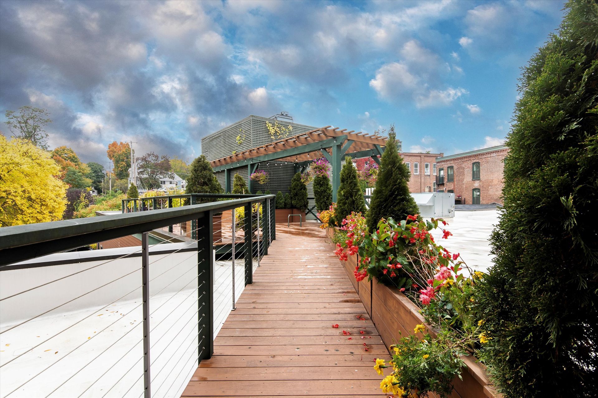 A wooden walkway with a black cable railing leads to a pergola on a rooftop terrace, lined with potted flowers and trees.